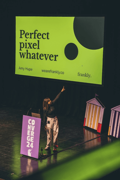 Amy Hupe pointing at the screen above her, displaying her slides during her Converge Brighton 2024 talk.